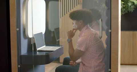 Young man working in glass booth with laptop, modern coworking private workspace
