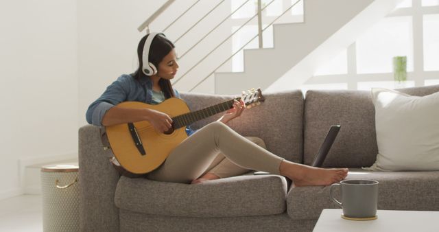 Image of happy biracial woman in headphones sitting on sofa and playing guitar. Lifestyle, music, hobby and spending free time at home concept.