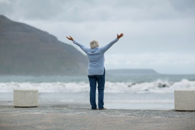 Senior woman standing with arms outstretched on beach, facing ocean waves. Ideal for themes of freedom, relaxation, retirement, and wellness. Suitable for travel brochures, wellness articles, and retirement planning materials.