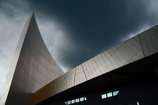 Showcasing a modern architectural structure with a minimalist design set against a dramatic sky, this image highlights angular and geometric shapes with a metal facade. Ideal for use in projects related to urban landscapes, contemporary architecture, city planning, and marketing materials for architectural firms.