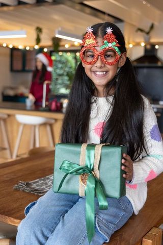 Smiling asian girl holding green gift wearing red christmas tree glasses in cozy kitchen
