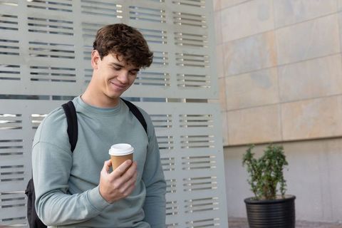 Smiling young man holding takeaway coffee cup beside urban metal screen with backpack