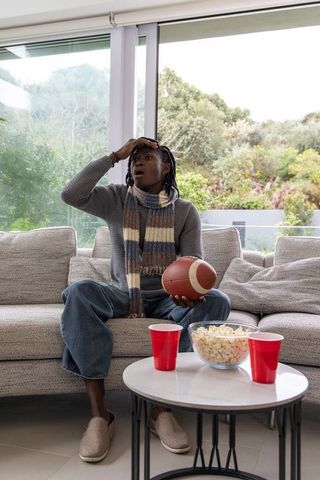African american woman reacting while holding football on couch with popcorn and red cups