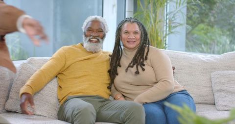 Senior couple sitting on sofa smiling and embracing in bright cozy living room with plants