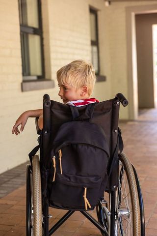 Child sitting in wheelchair with backpack looking left in school corridor
