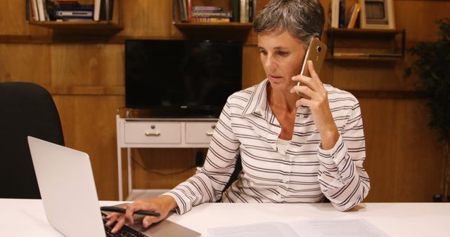 A mature woman is sitting at a desk in a home office, typing on a laptop while talking on a mobile phone. She is wearing a striped white shirt and appears to be focused on her work. Bookshelves and a television are visible in the background. This image is perfect for illustrating themes of remote work, productivity, modern business practices, and the use of technology in a home office environment.