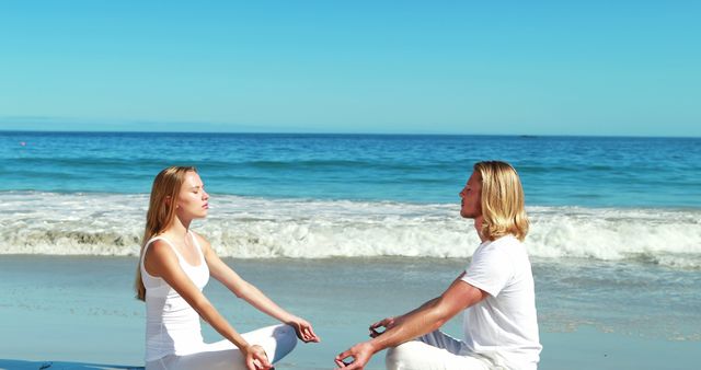 A young couple is meditating facing each other on a sandy beach with gentle ocean waves in the background. They are wearing white clothing, suggesting purity, peace, or a yoga session. Great for illustrating themes around mindfulness, wellness, outdoor fitness, relaxation techniques, and promoting healthy lifestyles. Could be used in wellness brochures, mental health awareness campaigns, yoga or fitness websites, and outdoor activity events.
