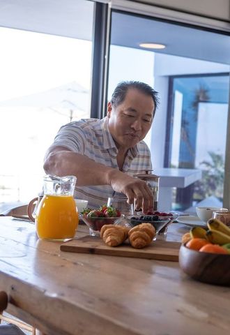 Mature chinese man enjoying sunlit terrace breakfast reaching fresh berries and croissant