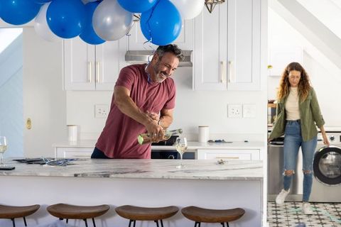 Man pouring wine into glass under blue silver balloons with partner entering kitchen