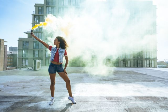 Front view of a hip young biracial woman wearing denim jacket and jeans, holding a smoke grenade with yellow smoke on an urban rooftop with building in the background.