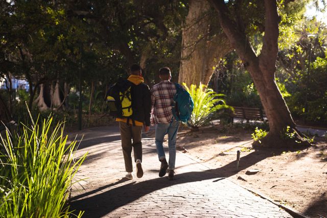 Two friends are walking along a path in a city park, each carrying a backpack. The scene is bathed in sunlight, with trees and greenery surrounding them. This image can be used for themes related to friendship, travel, outdoor activities, and leisure. It is ideal for promoting travel destinations, adventure tours, or lifestyle blogs.
