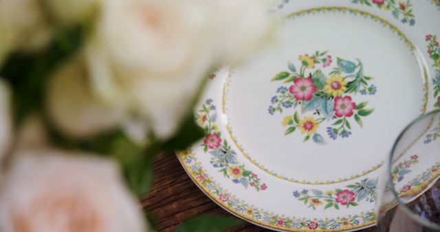 Elegant floral-patterned china plates are set on a wooden table, with a soft focus on a bouquet of roses in the foreground. The setting suggests a sophisticated event or a festive celebration, with a touch of vintage charm.