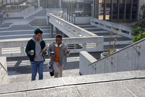 Two diverse students walking up campus stairs with coffee and notebooks in brutalist scene