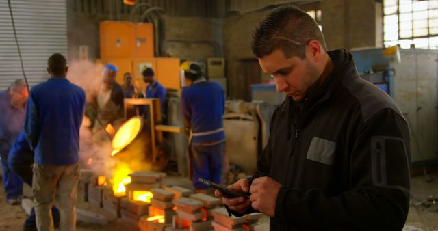 Engineer Using Smartphone in Foundry with Workers Smelting Metal - Download Free Stock Photos Pikwizard.com