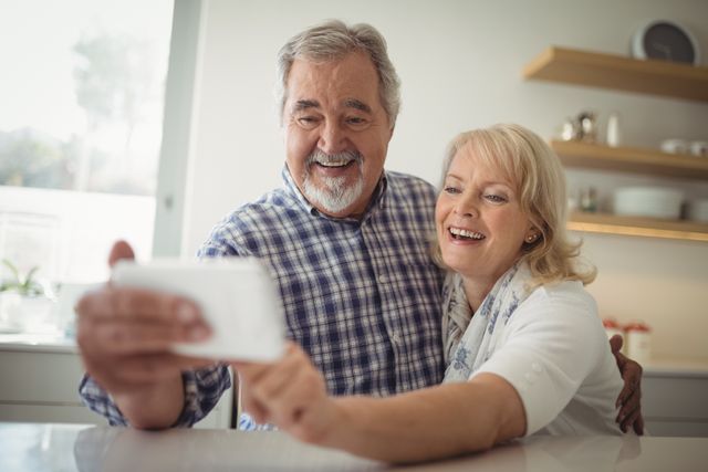 Senior couple smiling while taking a selfie in their modern kitchen, conveying happiness and togetherness. Ideal for advertisements about senior living, healthcare services, family bonds, and lifestyle blogs focusing on retirement and aging gracefully.