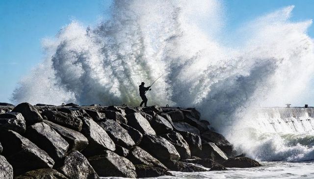 Silhouetted angler standing on rocky jetty facing crashing ocean waves and casting line