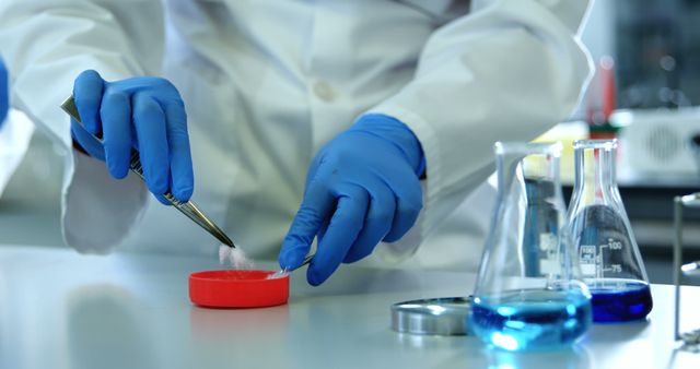 Scientist conducting laboratory experiment using various lab equipment and chemicals. Image shows a close-up of hands in blue gloves handling substances with lab tools. Ideal for illustrating concepts related to scientific research, chemistry studies, laboratory procedures, and educational materials about experiments.