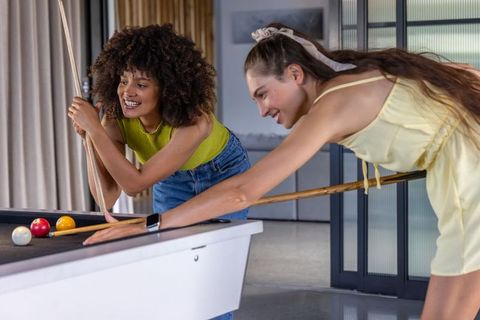 Multiracial friends playing pool and laughing in modern lounge wearing bright clothes