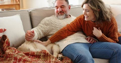 Smiling couple relaxing on sofa petting sleeping dog under cozy knit blanket