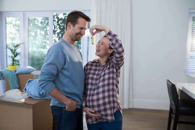 Pregnant couple smiling and holding key to their new house, surrounded by moving boxes. Ideal for use in real estate promotions, family lifestyle blogs, and advertisements for home services.