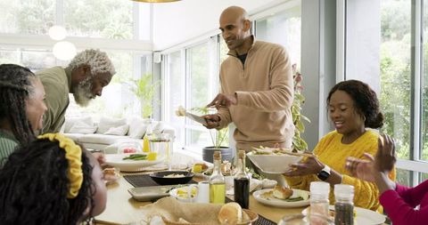 Multigenerational family sharing meal in sunlit modern dining room while passing plates
