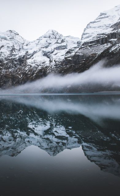 Snow-covered mountains reflecting in calm lake, with a layer of fog adding mystical ambiance. Perfect for nature-themed projects, travel brochures, winter and outdoor activities promotions, and tranquil scenery backgrounds.