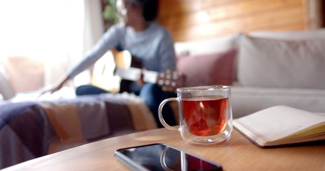 Glass cup of tea sits on wooden table next to smartphone and open notebook while person plays guitar in background. Ideal for themes of relaxation, home comfort, leisurely afternoons, or moments of creativity.
