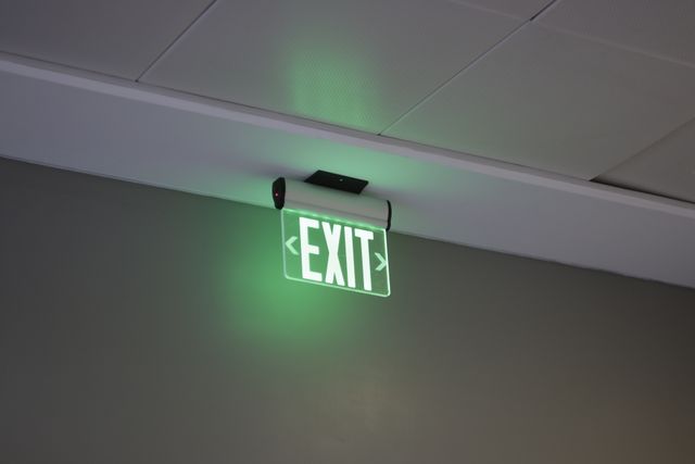 Illuminated green exit sign mounted on ceiling of a modern office building. Would be useful for articles or presentations on office safety, emergency evacuation plans, building regulations, or safe interior design.