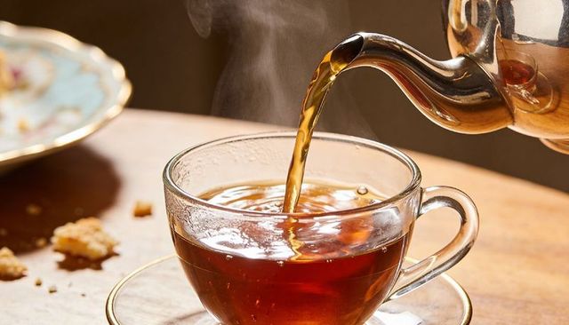 Pouring hot tea from polished silver teapot into clear glass teacup with steam on wooden table