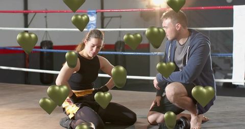 Female boxer resting on ring floor with coach kneeling and floating green heart overlays