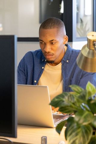 African american man focusing on laptop in modern office with greenery and brass lamp