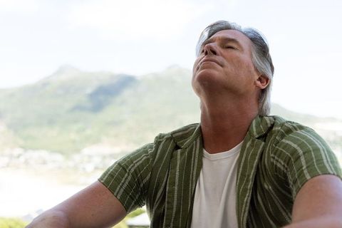 Middle-aged man gazing upward on balcony overlooking mountain view, relaxed contemplation