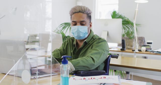 The image features a man in a wheelchair working at a desk in an office environment, wearing a protective face mask and typing on a computer. Hand sanitizer is placed nearby, indicating attention to hygiene and safety. This image is suitable for illustrating workplace inclusivity, health and safety measures during the pandemic, and professional work settings. It can be used in articles, blog posts, and marketing materials addressing disability support, workplace safety, and COVID-19 impacts on work environments.