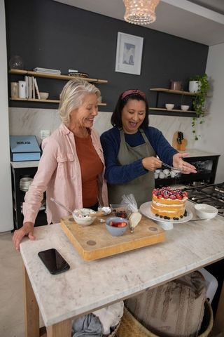 Senior friends decorating layered sponge cake with fresh berries in cozy home kitchen