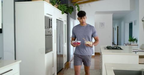 Young african american man carrying meal prep bowls with wooden spoon in modern kitchen