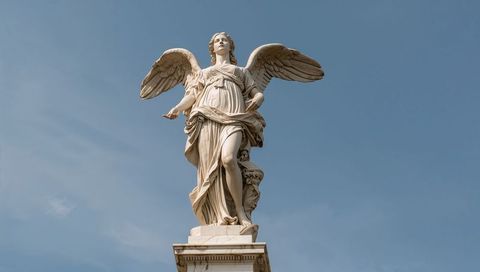 Marble angel statue standing on ornate pedestal with flowing drapery under pale blue sky