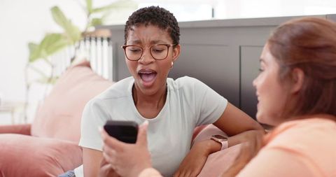 Surprised diverse friends reacting to smartphone while relaxing on pink sofa in sunlit living room