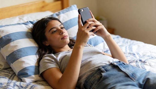 Teen girl reclining on bed scrolling smartphone in grey tee and jeans, relaxed at home