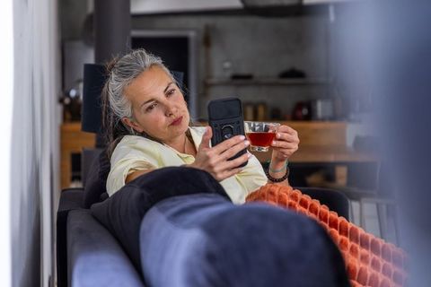 Senior woman reclining on sofa scrolling smartphone and sipping tea nestling in cozy throw