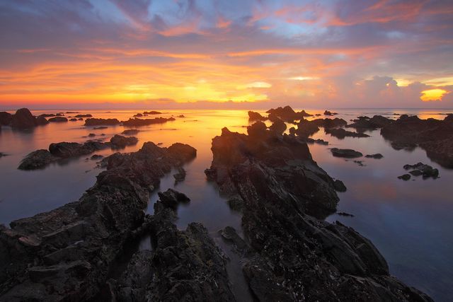 Perfect for use in travel brochures, nature magazines, and websites promoting serene destinations, this image captures the stunning colors of a sunset over a rocky seashore. The calm waters reflect the dramatic sky, creating a tranquil scene ideal for backgrounds and inspirational projects.