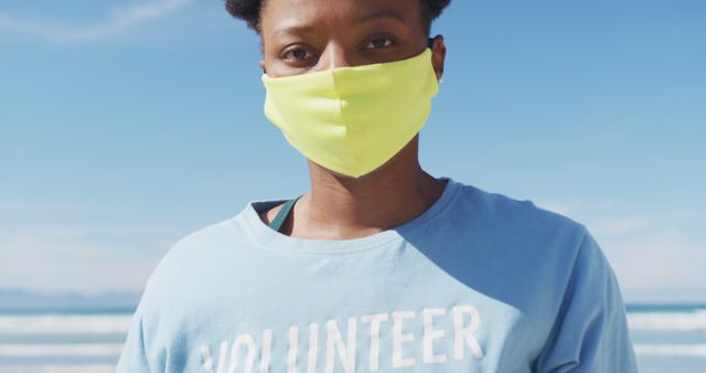 Portrait of african american woman wearing volunteer t shirt and face mask looking at camera. eco conservation volunteers, beach clean-up during coronavirus covid 19 pandemic.