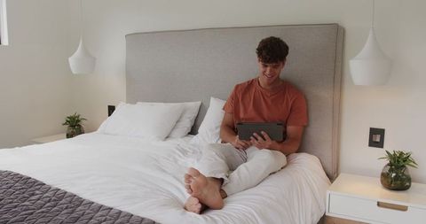 Young man sitting on bed using tablet in bright minimalist bedroom for remote leisure