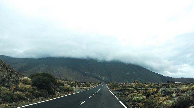 The image depicts an empty asphalt road leading towards a mountain range shrouded in fog and clouds. Surrounding the road are sparse, dry bushes and rocky terrain. This image is ideal for depicting themes of travel, adventure, exploration, and scenic landscapes. It can be used in travel blogs, nature magazines, promotional materials for outdoor activities, and websites focusing on road trips or journeys.