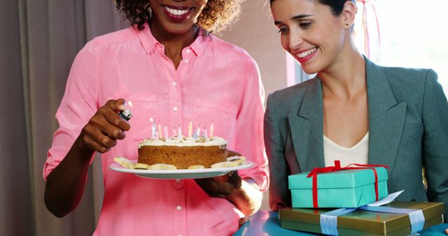 Two female colleagues celebrating a birthday at work; one is holding a cake with lit candles while the other holds wrapped presents. Ideal for illustrating corporate events, team building, office culture, and celebrations in professional settings.