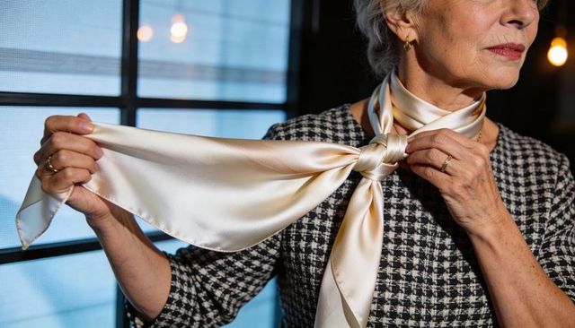 Senior woman tying cream silk scarf over houndstooth dress, closeup of hands and jewelry