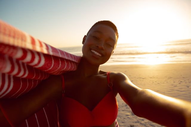 Woman standing on beach at sunset, holding a blanket and smiling. Ideal for use in travel brochures, lifestyle blogs, summer vacation promotions, and advertisements focusing on relaxation and outdoor activities.