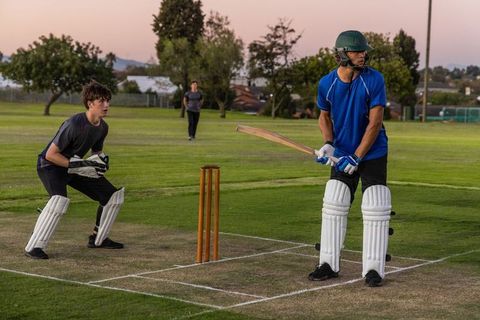 Evening cricket practice batting and wicketkeeping on grass pitch at dusk