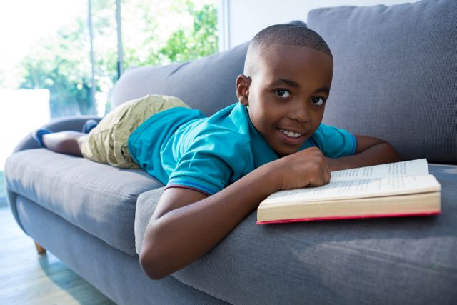 Young boy lying on a sofa in a living room, enjoying a book. Ideal for themes related to childhood, education, leisure, and home life. Can be used in educational materials, advertisements for home furnishings, or articles about reading habits in children.