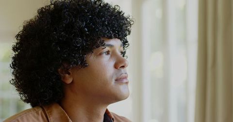 Curly-haired man reflecting by sunlit window indoors