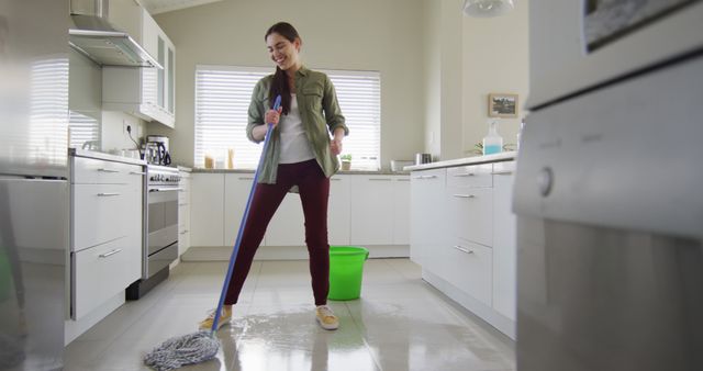 Young woman is mopping a clean, white kitchen floor, enjoying her housekeeping task. She smiles as she holds the mop with a green bucket nearby. Ideal for demonstrating household cleaning, home maintenance, domestic chores, or advertisements for cleaning products. Useful for lifestyle blogs, housekeeping articles, or cleaning service promotions.